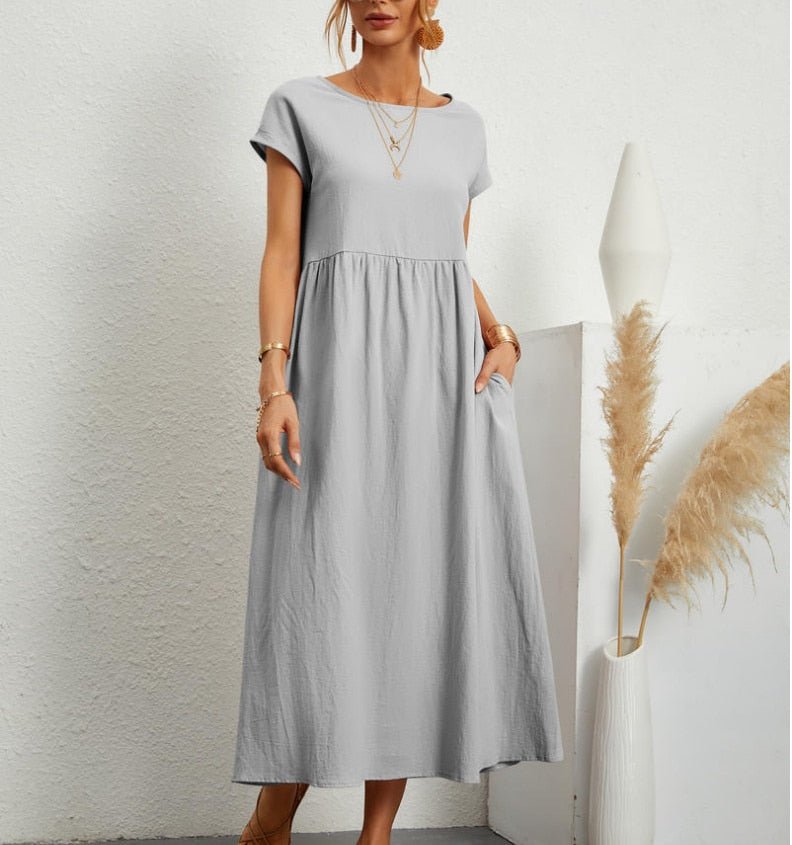 Frau in einem dunkelgrünen Sommerkleid hält eine Tasche und steht vor einer weißen Wand.